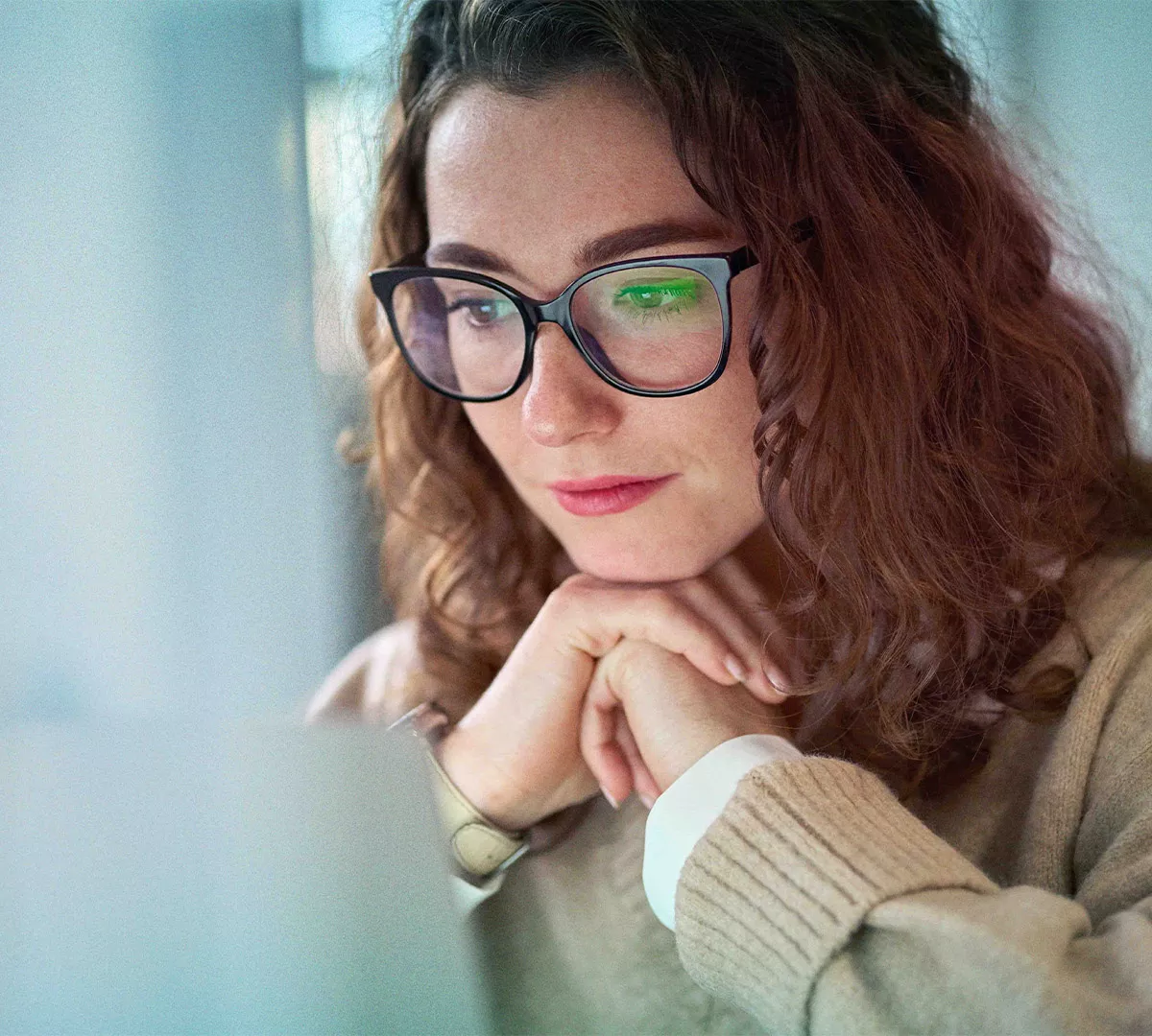 A person wearing a beige sweater and glasses is seated indoors, resting their hands together. The setting appears calm and reflective, with soft natural lighting enhancing the cozy atmosphere. The focus is on the hands and attire, with a blurred background adding depth.